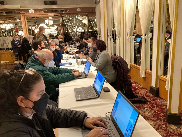 Inside a hotel conference room, people sit at tables at laptops typing in